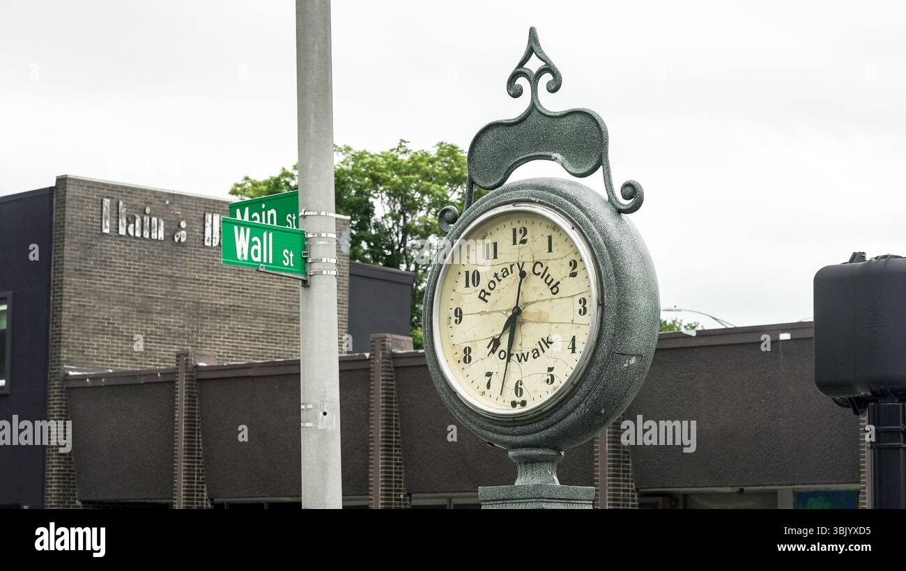 NORWALK, Connecticut, USA - 16 GIUGNO 2025: Un orologio d'epoca si erge in primo piano all'incrocio tra Main Street e Wall Street. Ombreggiato da cieli sovrastati, il Foto Stock