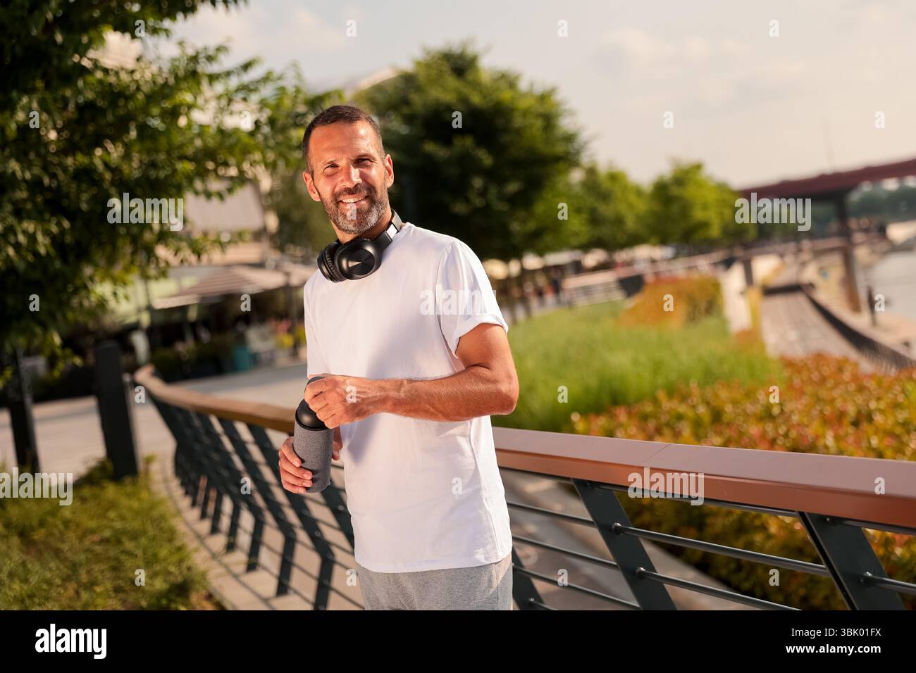 Un uomo allegro sta in piedi vicino all'acqua, crogiolandosi alla luce del sole con una bevanda calda in mano, cuffie intorno al collo, godendosi l'atmosfera serena. Foto Stock
