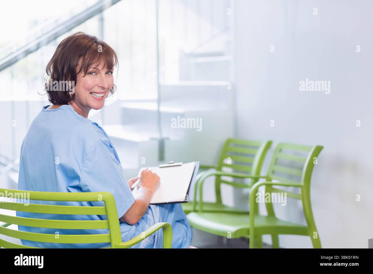 Infermiera donna seduta su una sedia verde nella sala d'attesa, che scrive sugli appunti sorridente, copia dello spazio Foto Stock