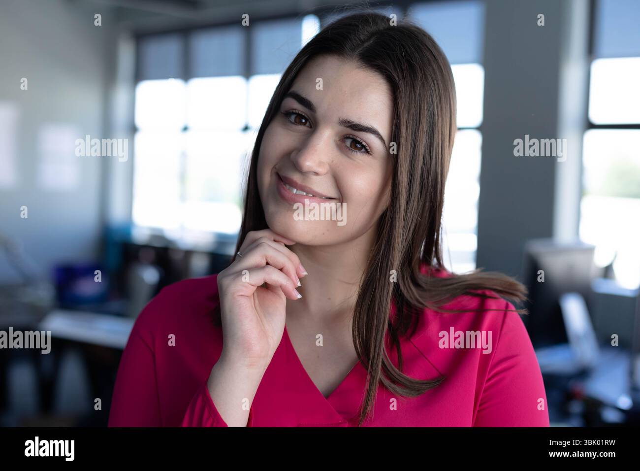 Donna che riposa il mento in un ufficio open space con camicetta rosa con tende alla finestra e monitor per computer Foto Stock