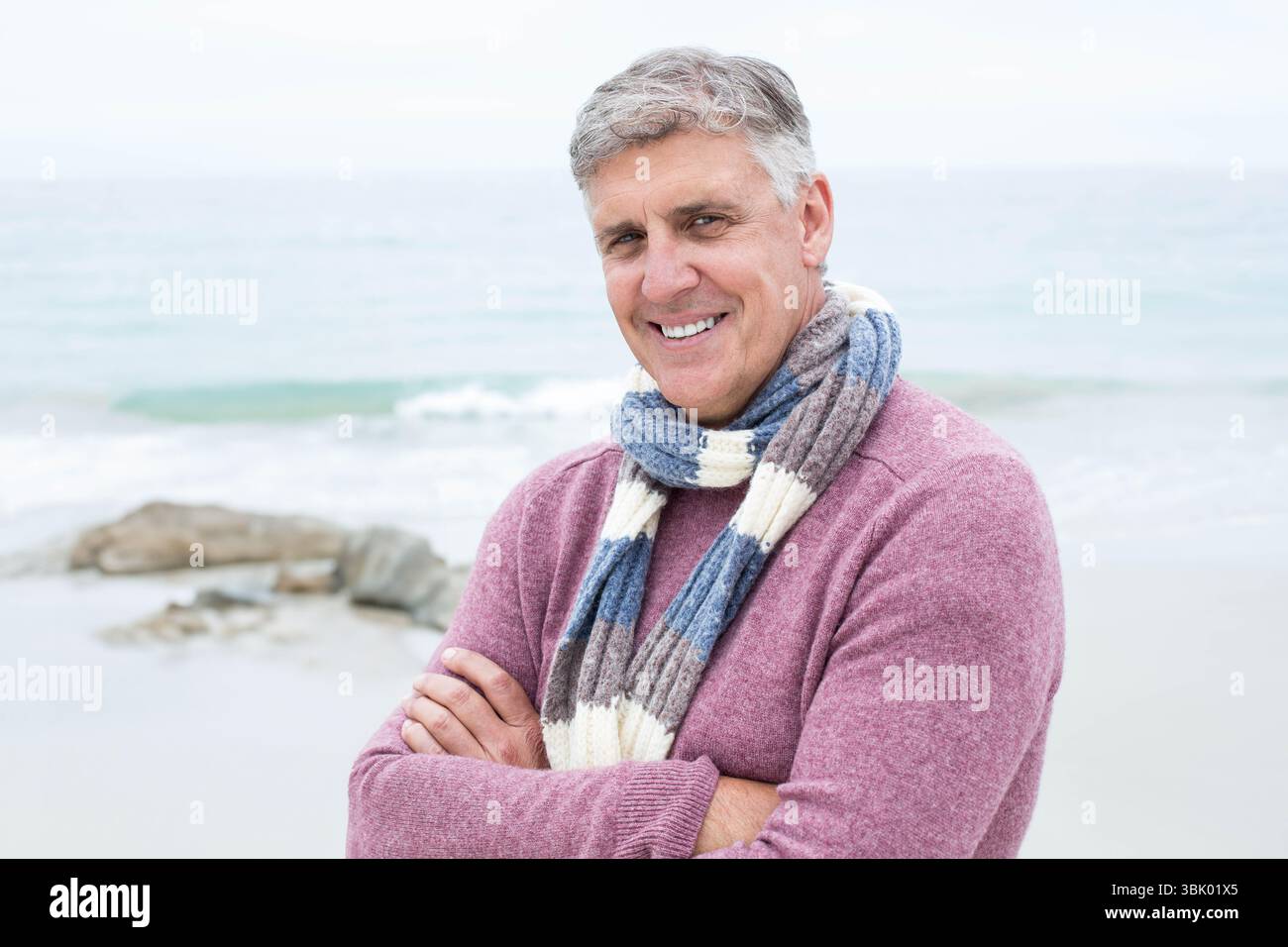 Uomo anziano in piedi sulla spiaggia sabbiosa con rocce costiere che indossano un maglione e una sciarpa a righe che sorride Foto Stock