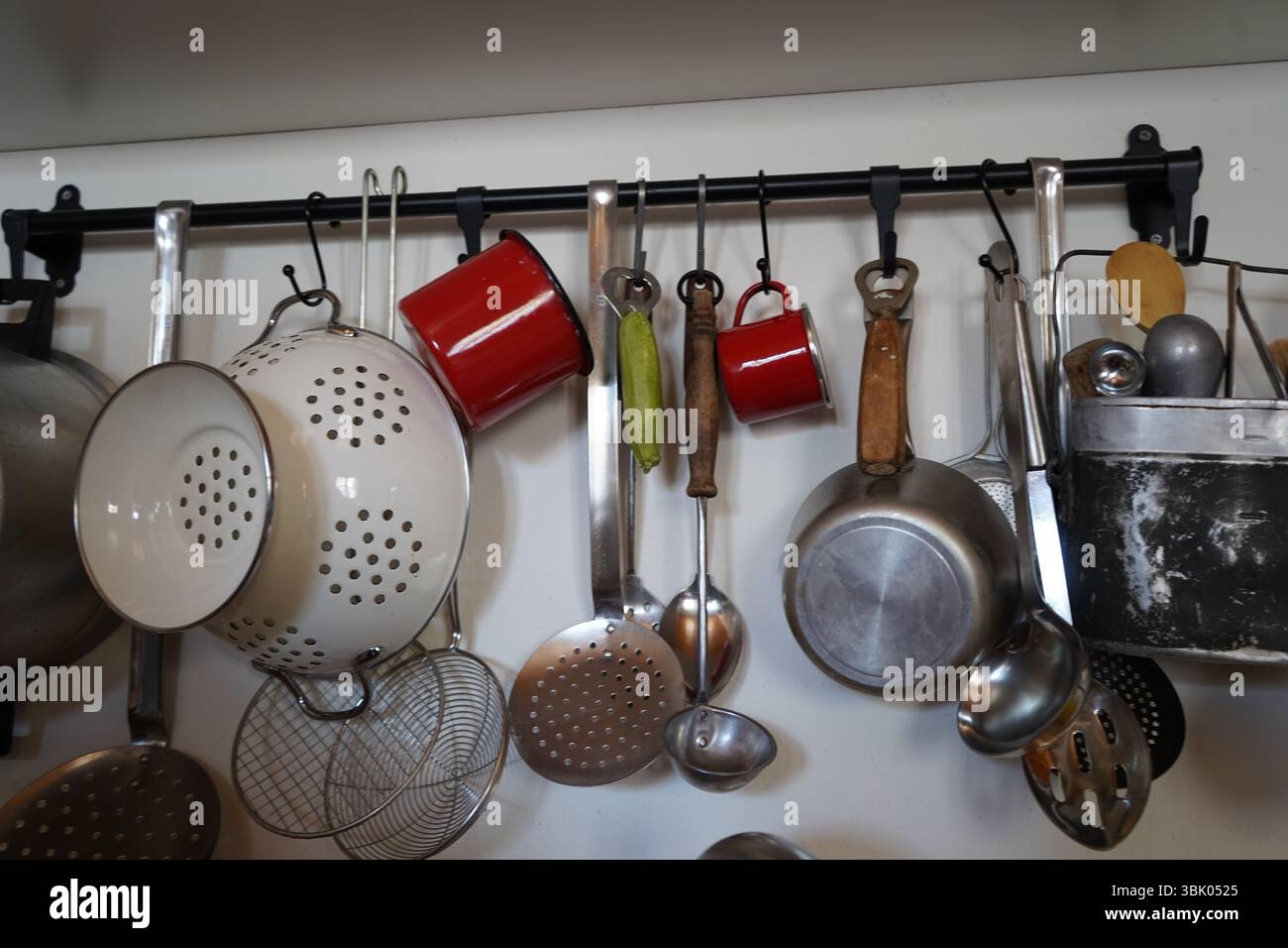 Una varietà di utensili da cucina è appesa a un'asta montata a parete. Gli articoli includono un colino bianco, tazze rosse, filtri metallici, mestoli e spatole. Foto Stock