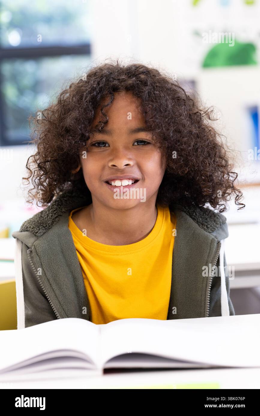 Ragazzo sorridente in classe che si diverte a imparare con un libro aperto sulla scrivania, a scuola Foto Stock