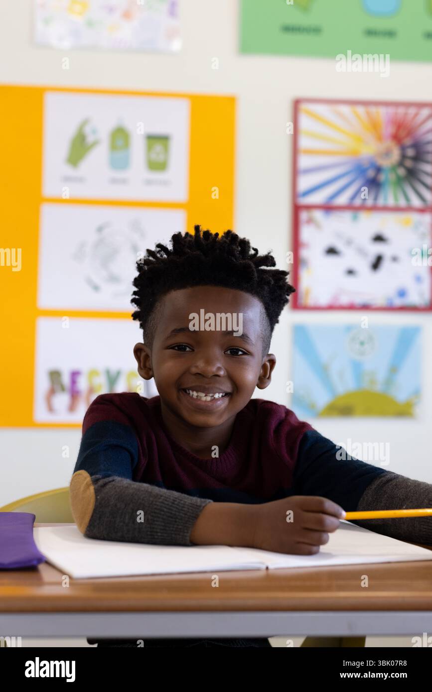 Sorridente ragazzo afroamericano a scuola che tiene in mano una matita, seduto alla scrivania con un notebook aperto Foto Stock