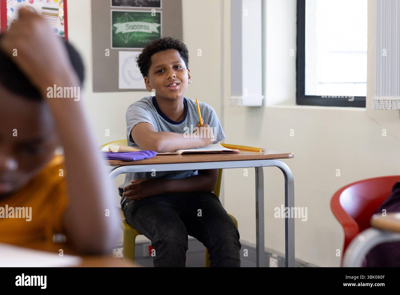 In classe, ragazzo che tiene in mano una matita e sorride mentre seduto alla scrivania, a scuola Foto Stock