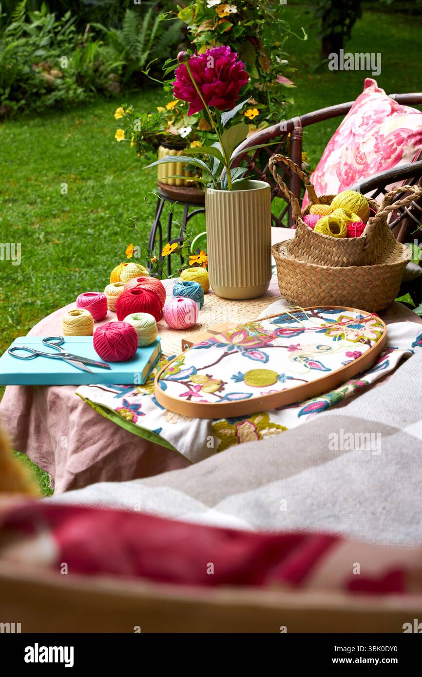 Primo piano di un piccolo tavolo da giardino con ricami in una calda giornata estiva. Fili colorati, aghi, tessuti, forbici e un cerchio in legno Foto Stock