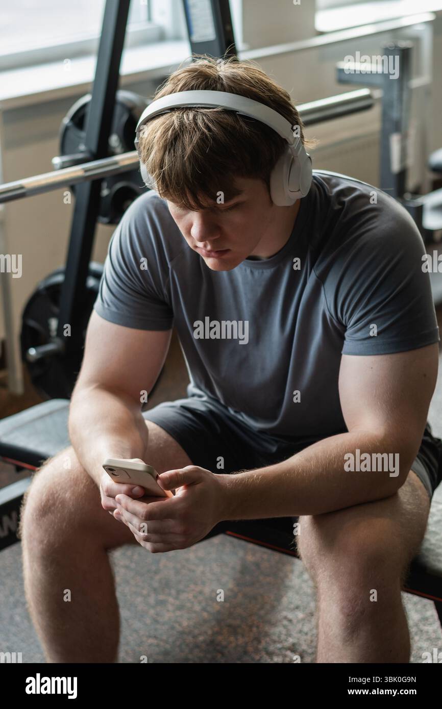 Giovane uomo sportivo caucasico rilassarsi e sedersi con il telefono cellulare, ascoltare musica cuffie e messaggi in palestra. Allestitore durante la formazione. Fitness healt Foto Stock