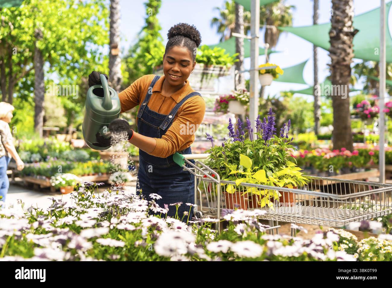 Giovane donna felice che lavora in un centro giardino annaffiando piante con una lattina mentre spinge un carrello della spesa Foto Stock