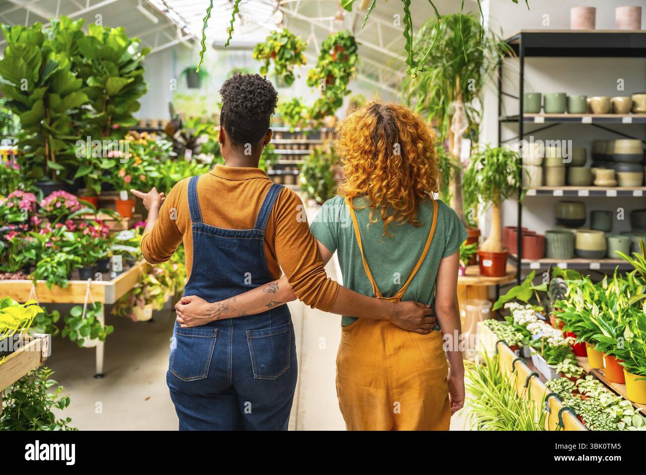 Due giovani donne che lavorano in un centro giardino camminano e discutono di piante in vendita, viste da dietro Foto Stock