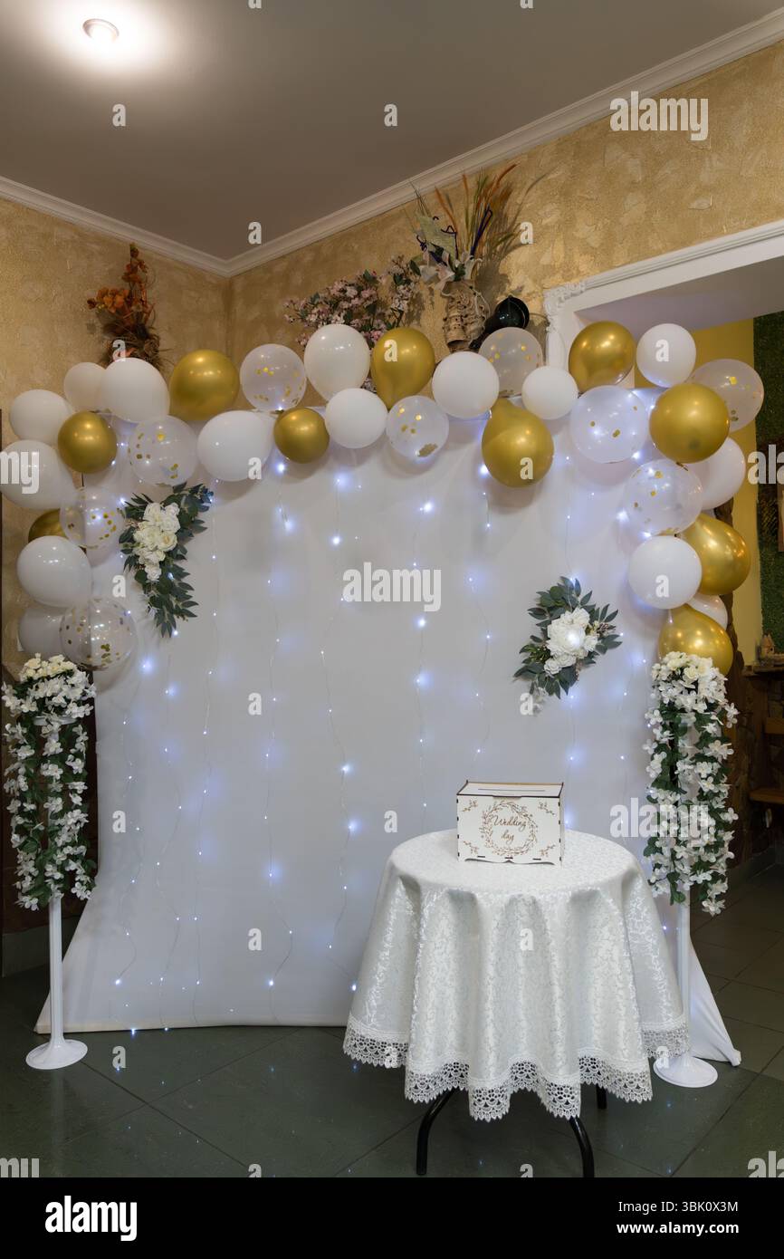 Uno splendido sfondo bianco per matrimoni è decorato con una ghirlanda in mongolfiera, accenti dorati, composizioni floreali a cascata, un tavolo da libro per gli ospiti e una calda ligh Foto Stock