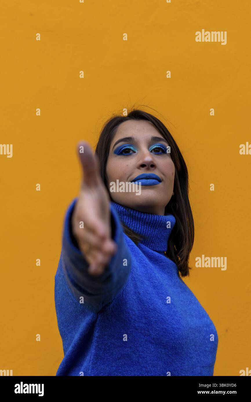 Ritratto verticale di donna caucasica con trucco blu e vestito di blu alzando la mano per stringere le mani su uno sfondo giallo Foto Stock