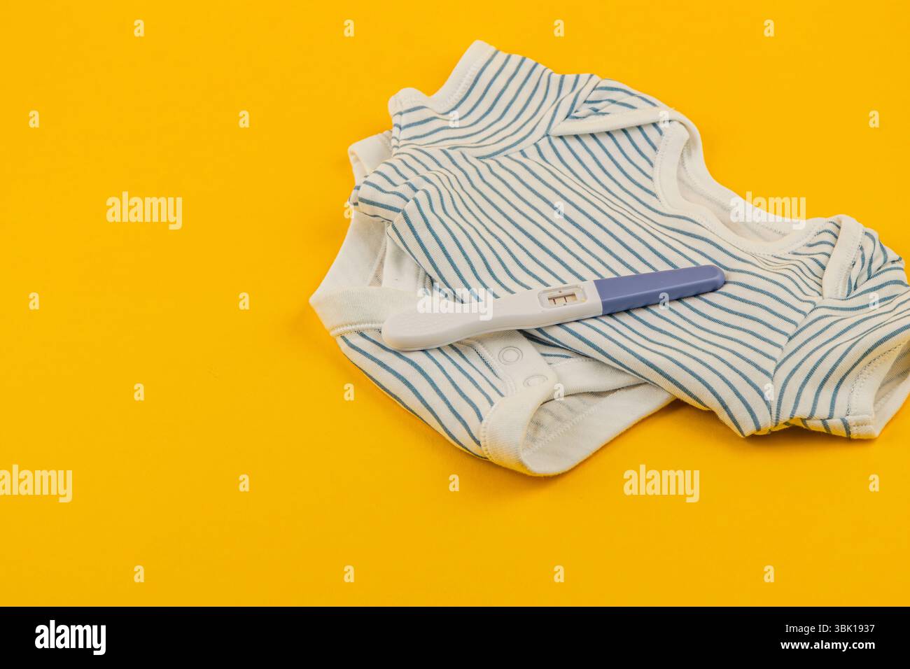 Gli accessori per neonati a righe sono impilati in modo ordinato accanto a un test di gravidanza su una superficie gialla vibrante, simboleggiando nuovi inizi e la maternità. Foto Stock