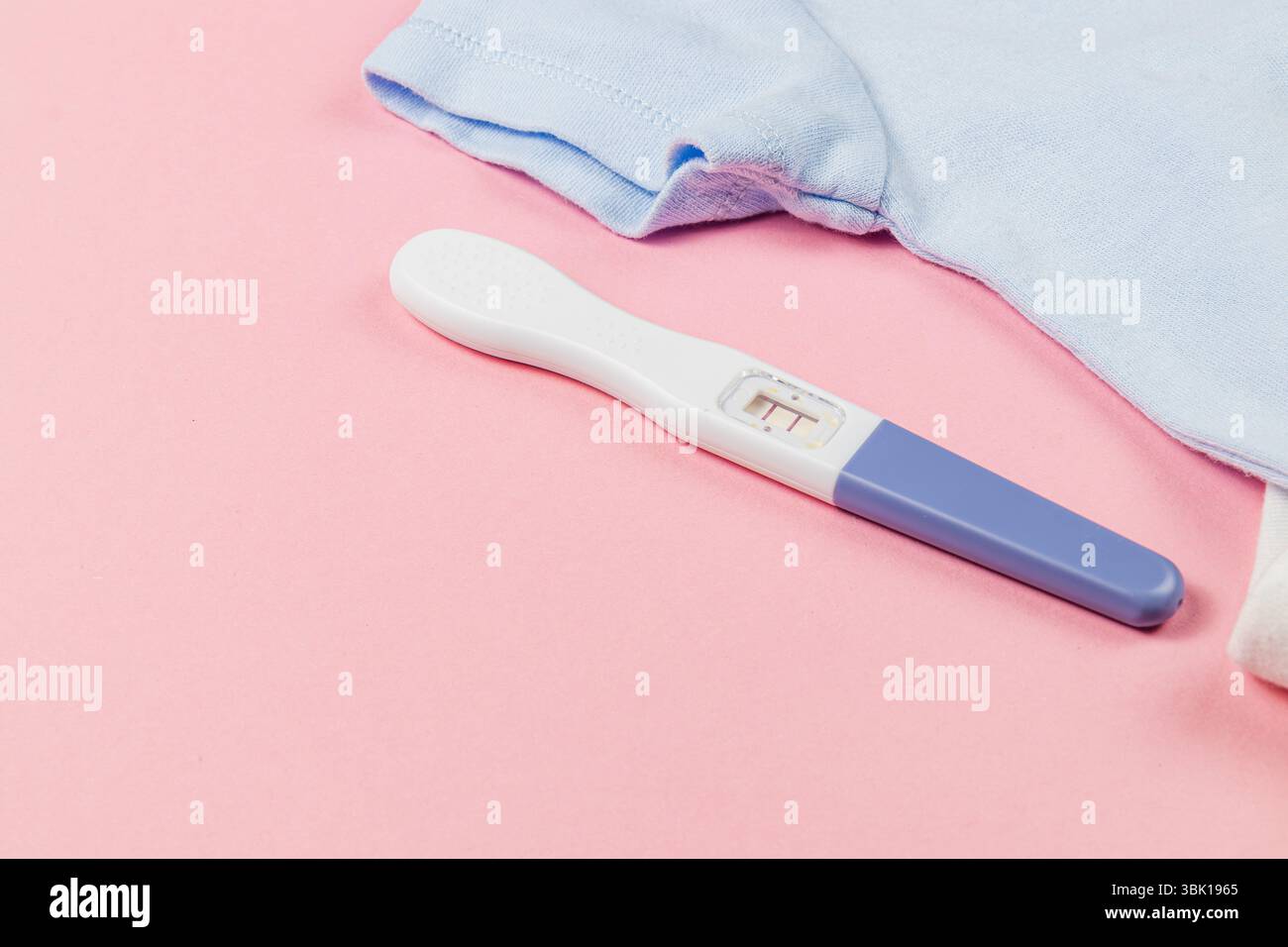 Un test di gravidanza mostra un risultato positivo insieme a un bel vestito blu per bambini su una superficie rosa tenue, simboleggiando nuovi inizi e anticipazioni. Foto Stock