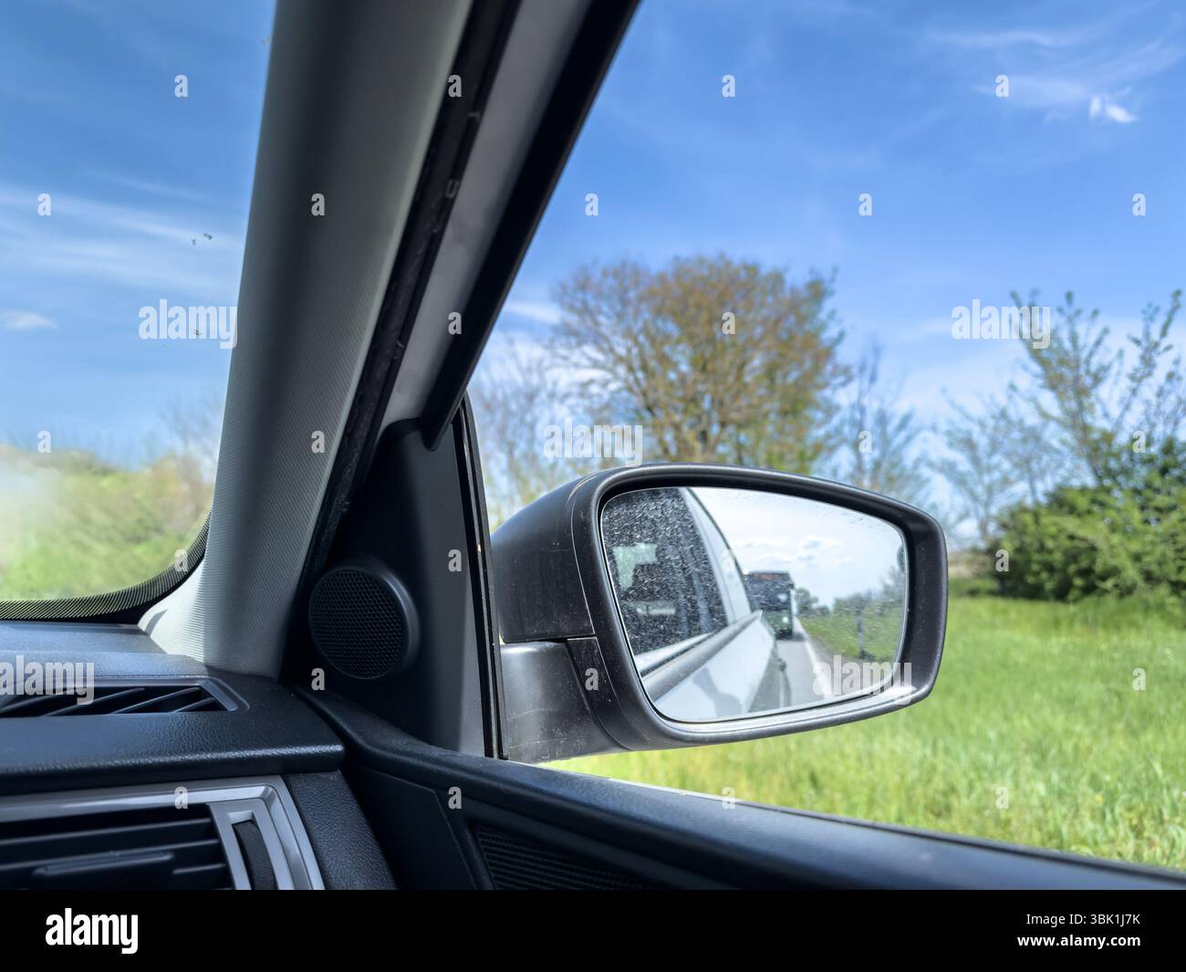 La vista lato conducente cattura uno specchio polveroso che riflette un autobus in avvicinamento da dietro su una strada rurale a due corsie, incorniciata da alberi verdi e primaverili sotto Foto Stock