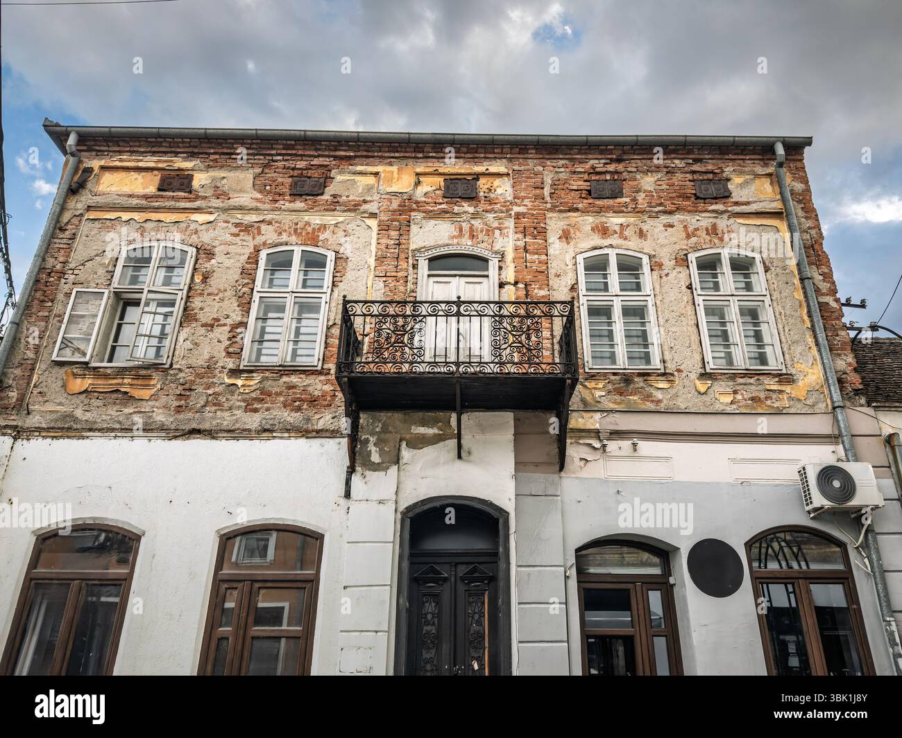 Residenza in mattoni del XIX secolo a due piani nella via centrale di Valjevo mostra intonaco che si stacca, balcone arrugginito e riparazioni delle finestre non corrispondenti sotto una tempesta Foto Stock