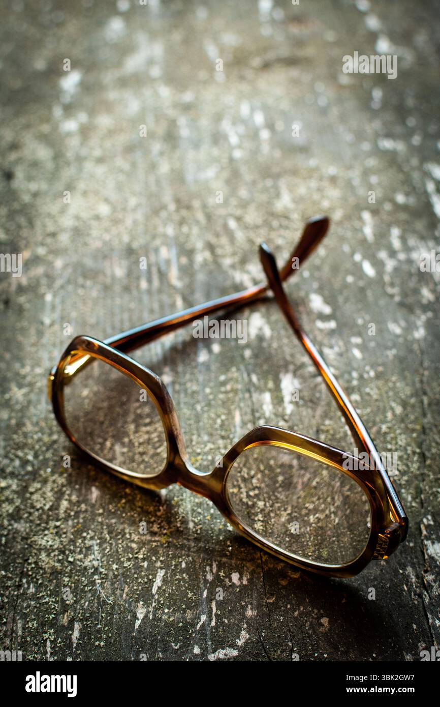 Un paio di occhiali vintage giace su una superficie in legno resistente agli agenti atmosferici, impreziosita da una calda luce naturale che si riflette sulle delicate lenti Foto Stock