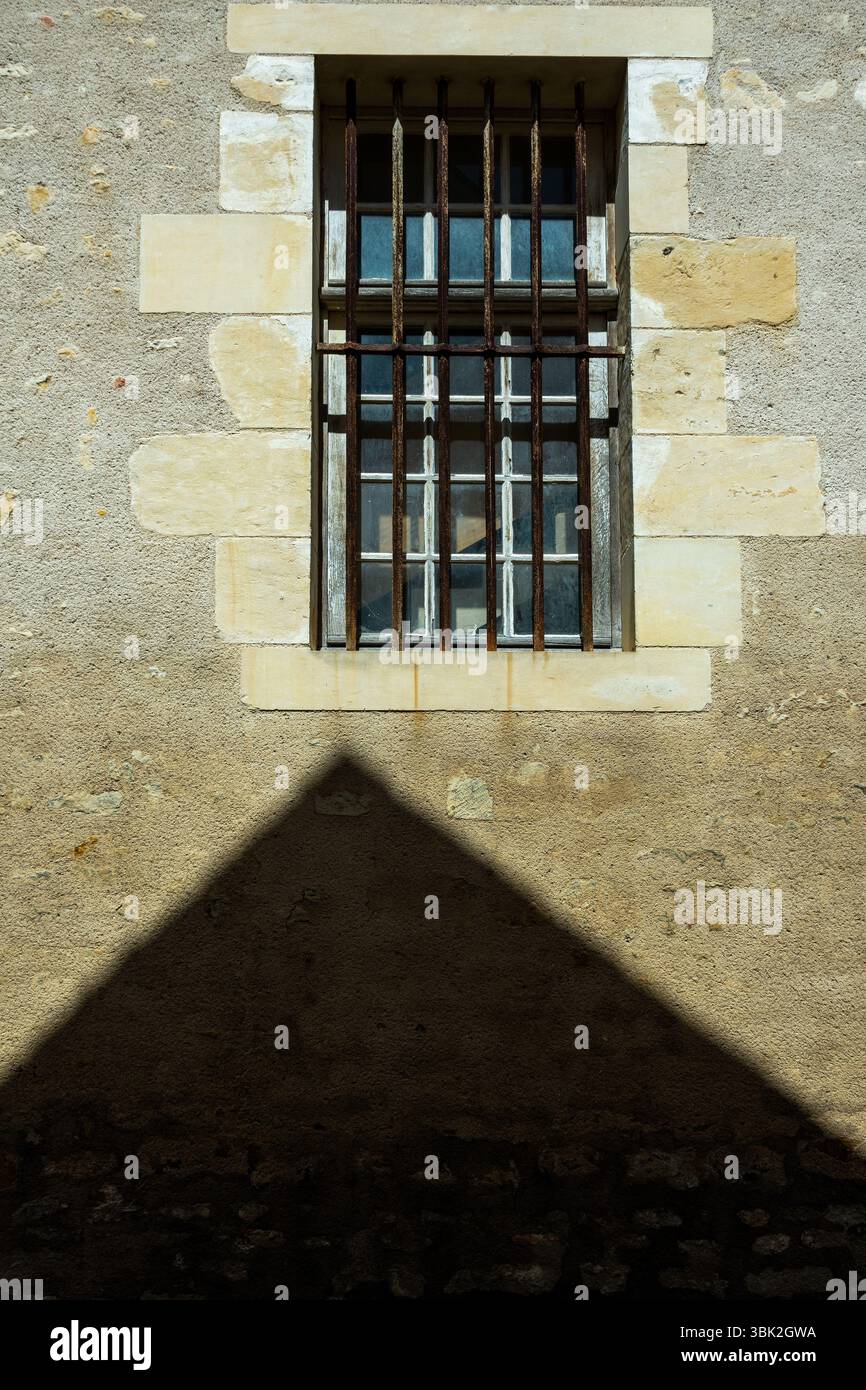 Una finestra sbarrata posizionata su un muro di pietra ruvida getta una forte ombra sotto. Francia Foto Stock