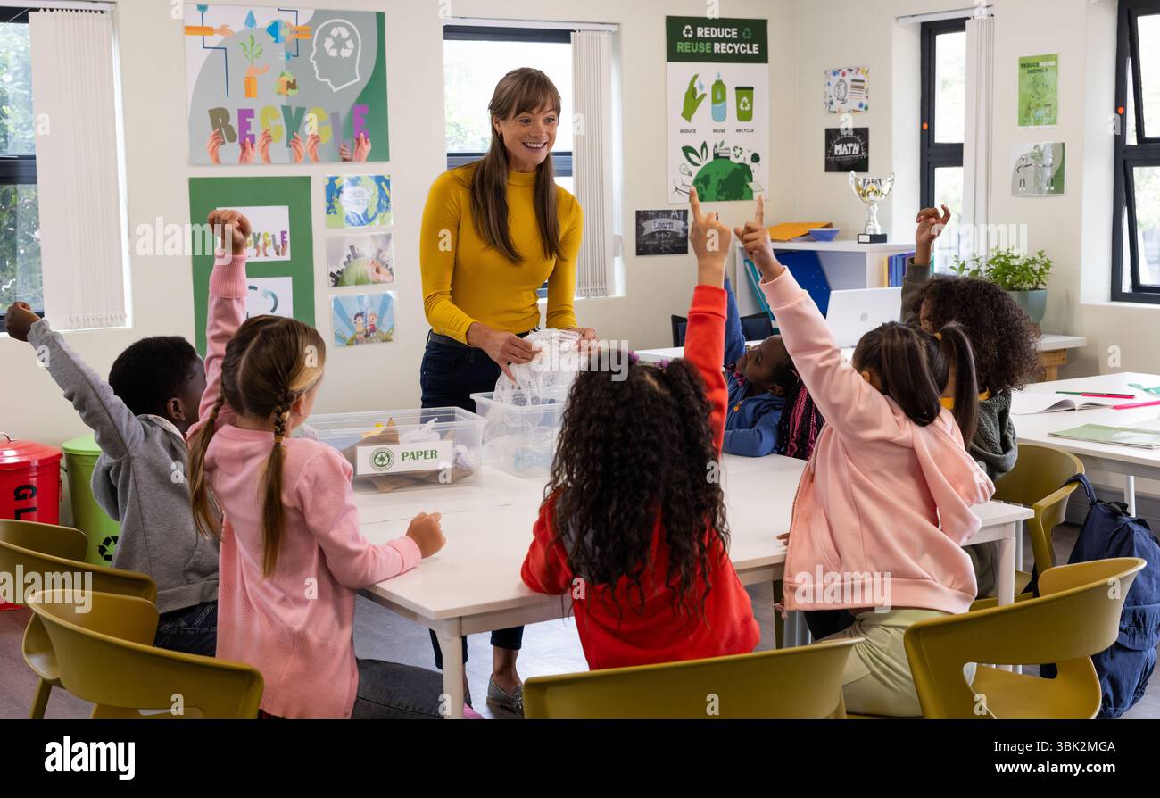 A scuola, insegnante femminile che coinvolge studenti diversi nella lezione di riciclaggio, alzando le mani Foto Stock