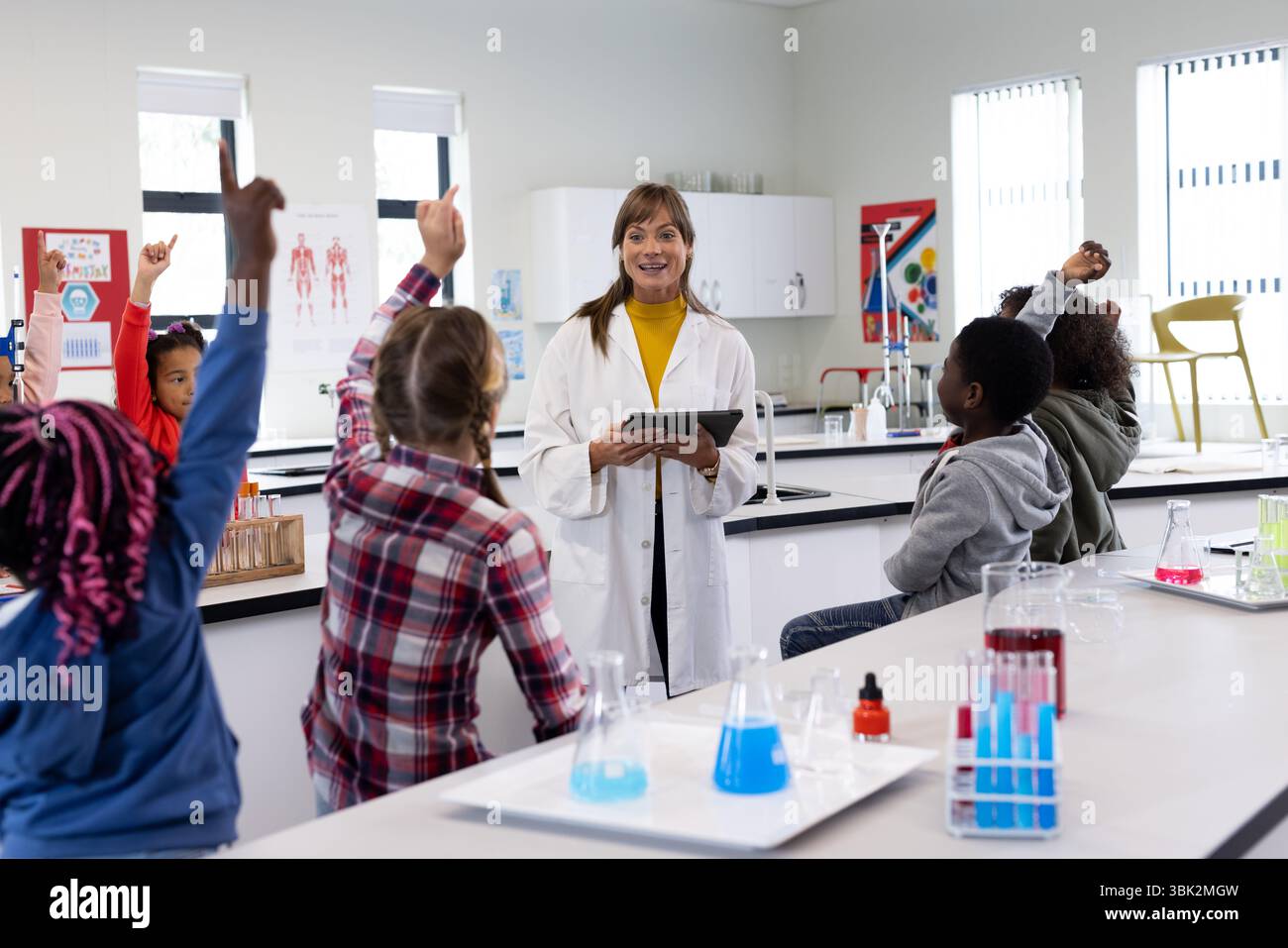 A scuola, insegnante femminile che coinvolge studenti diversi in classe di scienza, bambini che alzano le mani con impazienza Foto Stock