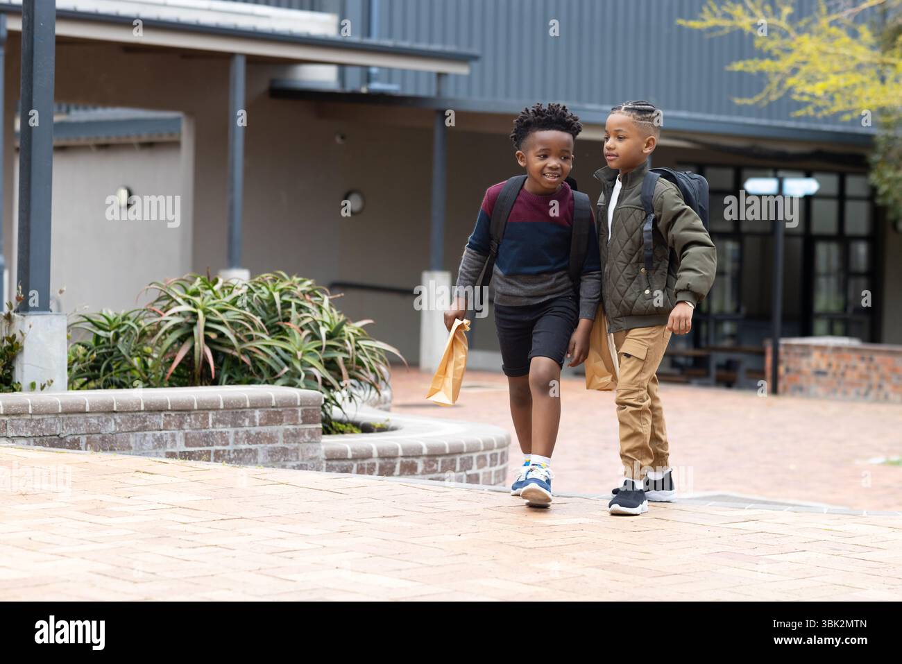 Due ragazzi diversi che camminano fuori scuola, trasportano zaini e sacchetti per il pranzo, sorridendo insieme Foto Stock