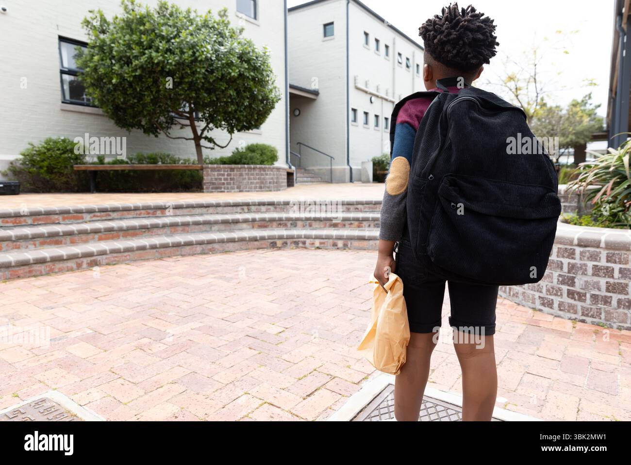 Ragazzo afroamericano con zaino che cammina verso l'edificio scolastico, tenendo in mano il cestino per il pranzo Foto Stock