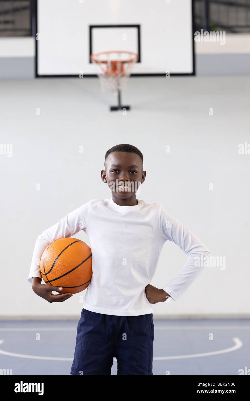 Sorridente ragazzo afro-americano che tiene il basket nella palestra scolastica, pronto per l'allenamento Foto Stock