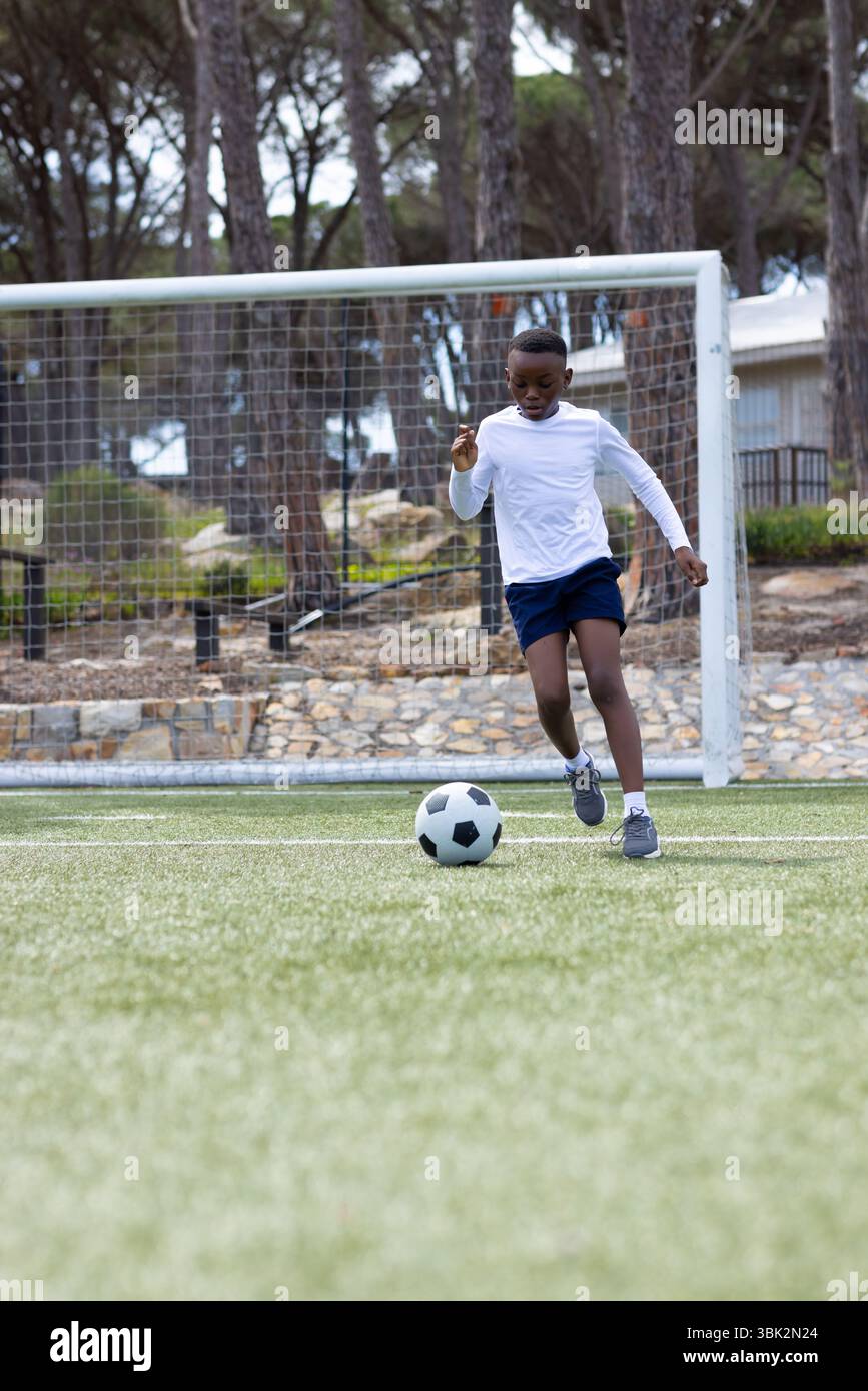 Ragazzo afroamericano che gioca a calcio sul campo, concentrandosi sul dribbling della palla verso la porta Foto Stock