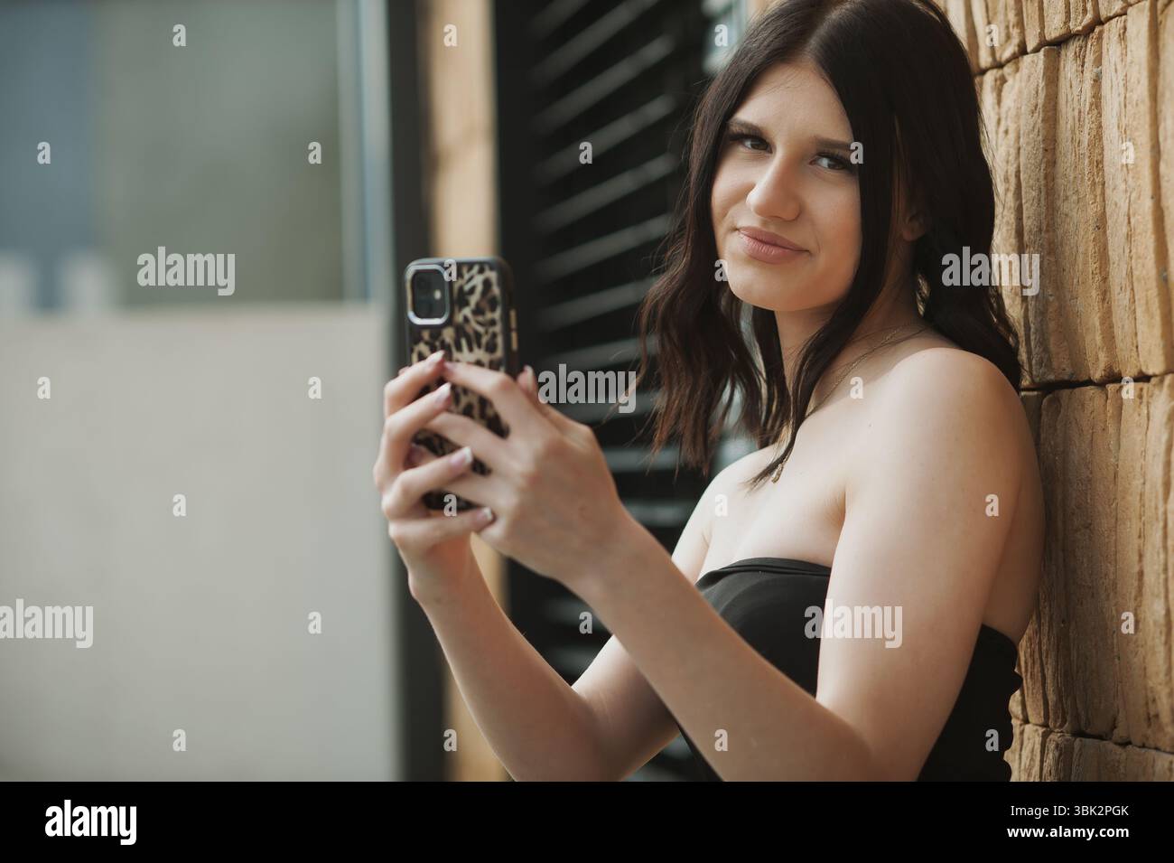 Una giovane donna con capelli castani di media lunghezza sta fuori, sorridendo mentre scatta un selfie. Lo sfondo presenta una parete testurizzata e una luce solare brillante Foto Stock
