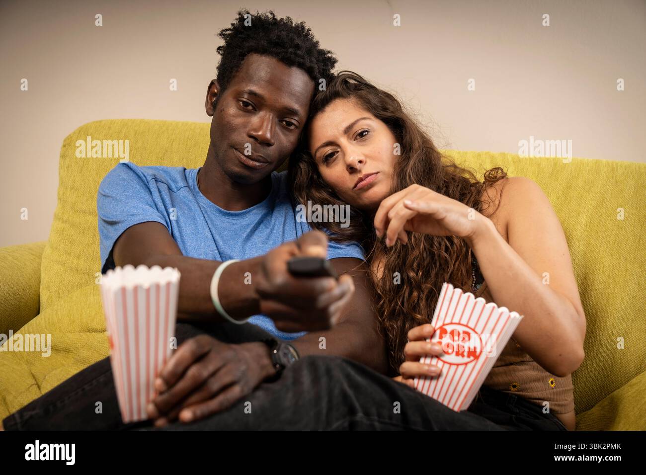 Una coppia giovane e variegata si siede sul divano per una serata al cinema a casa, con l'uomo che punta il telecomando direttamente verso la fotocamera, come se scegliesse una m Foto Stock