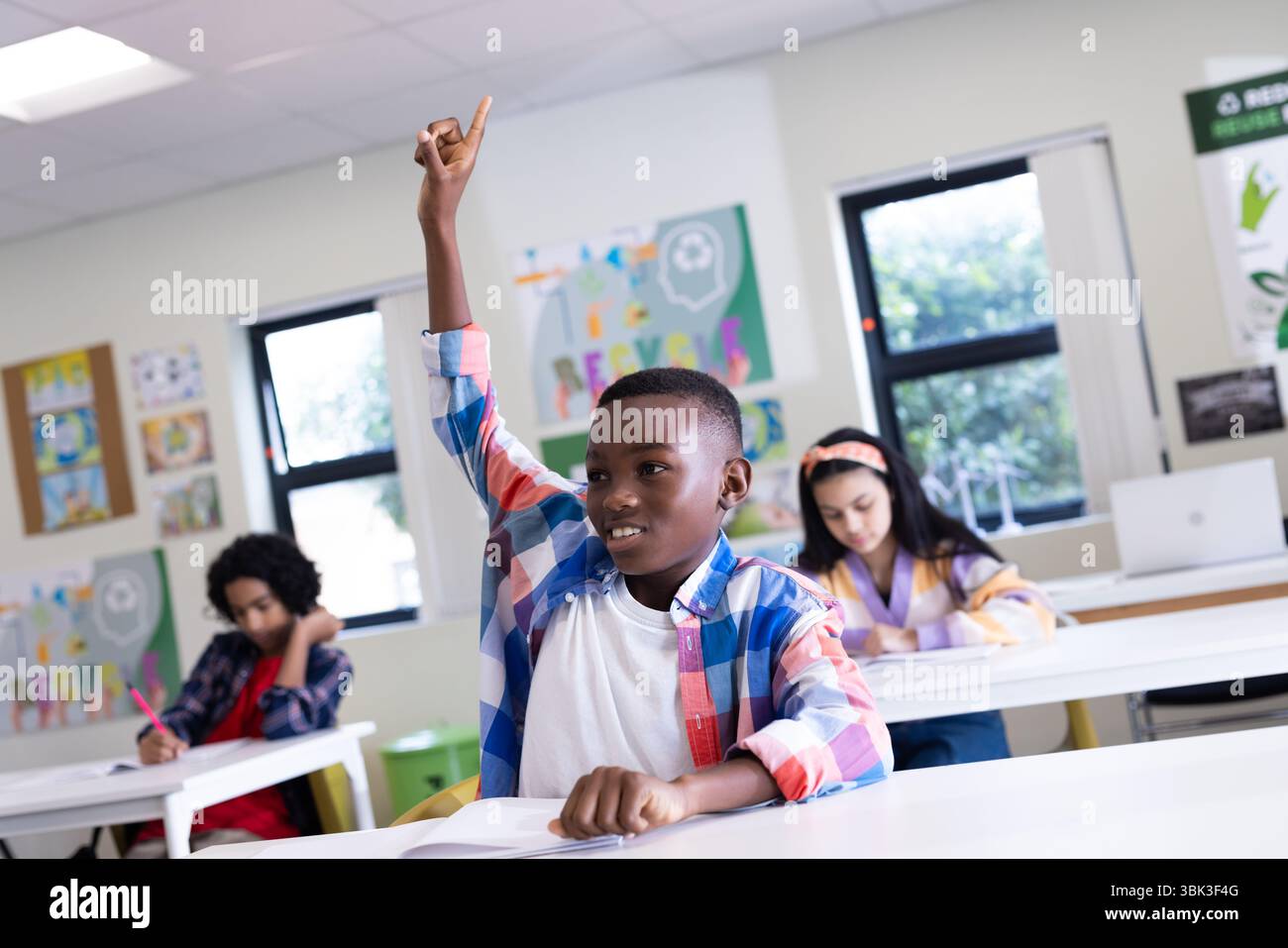 In classe, ragazzo che alza la mano mentre i compagni di classe scrivono, impegnato nell'apprendimento, a scuola Foto Stock