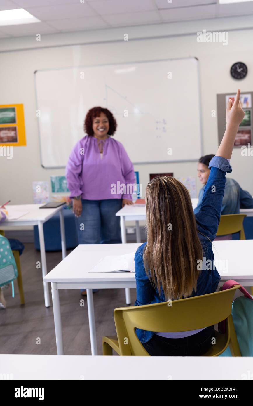 Ragazza che alza la mano in classe mentre la maestra sorride, si impegna in lezione, a scuola Foto Stock