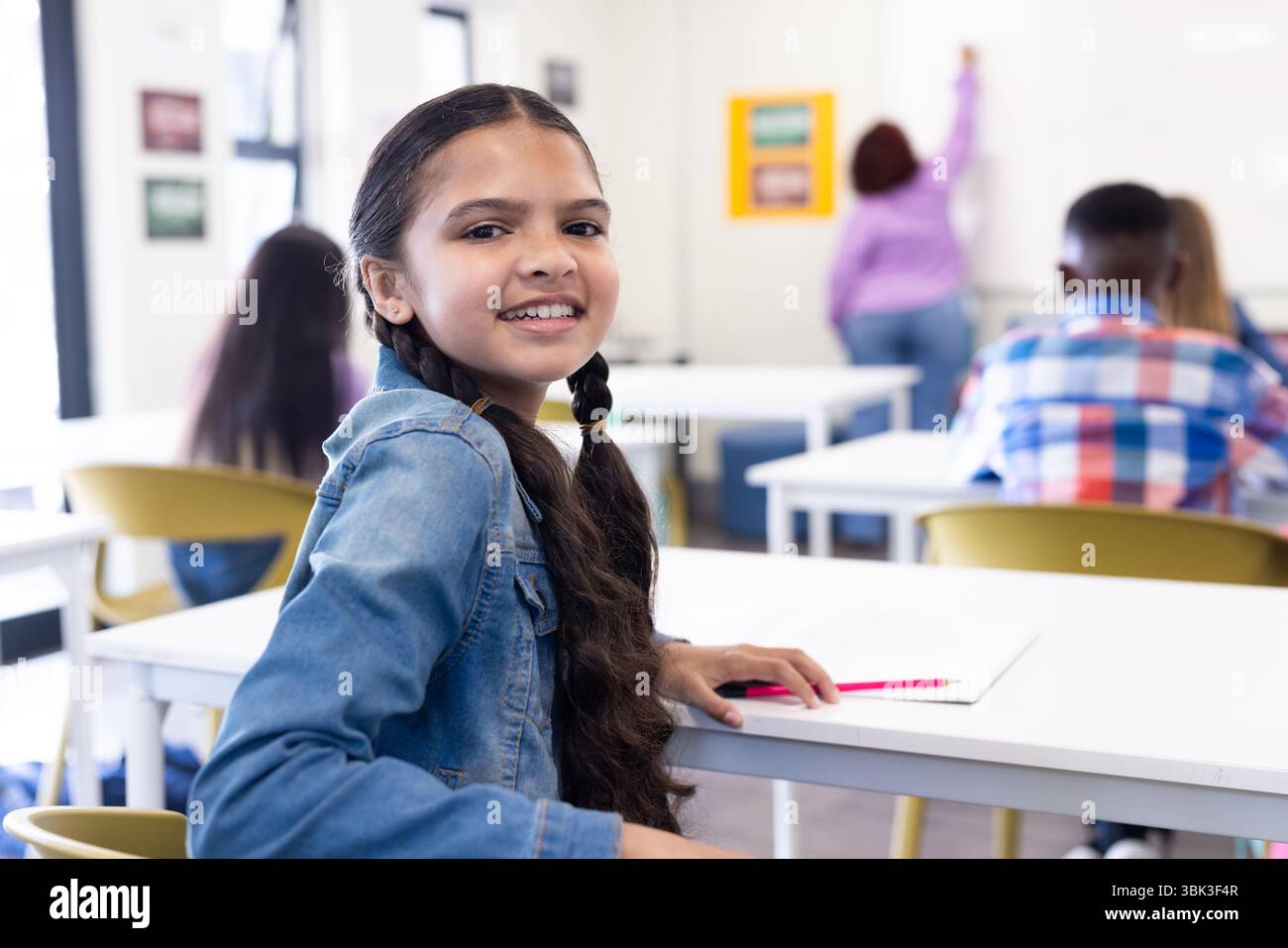 Ragazza sorridente in classe che guarda indietro, che tiene in mano la matita, insegnante donna che scrive a bordo, a scuola Foto Stock