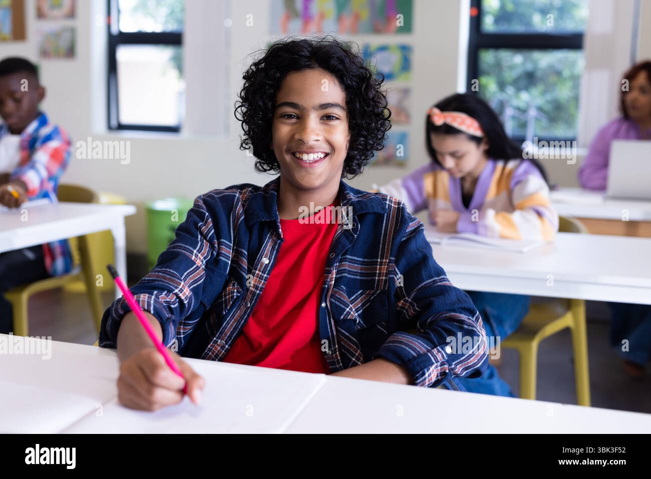 A scuola, ragazzo sorridente che scrive in classe, impara con i compagni di classe Foto Stock