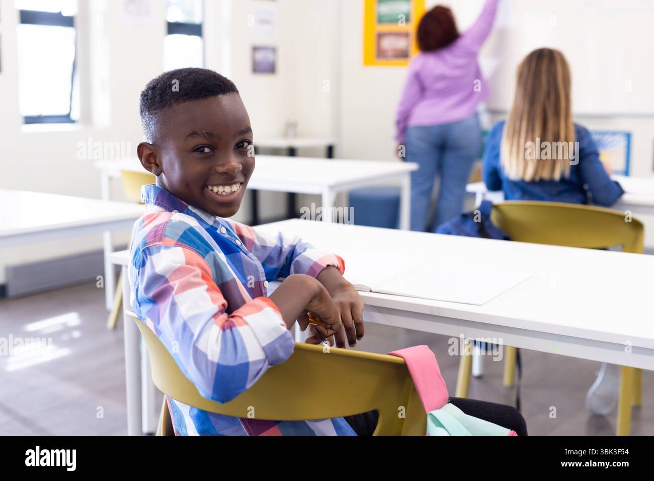 Un ragazzo sorridente in classe seduto alla scrivania mentre un'insegnante donna scrive a bordo, a scuola Foto Stock