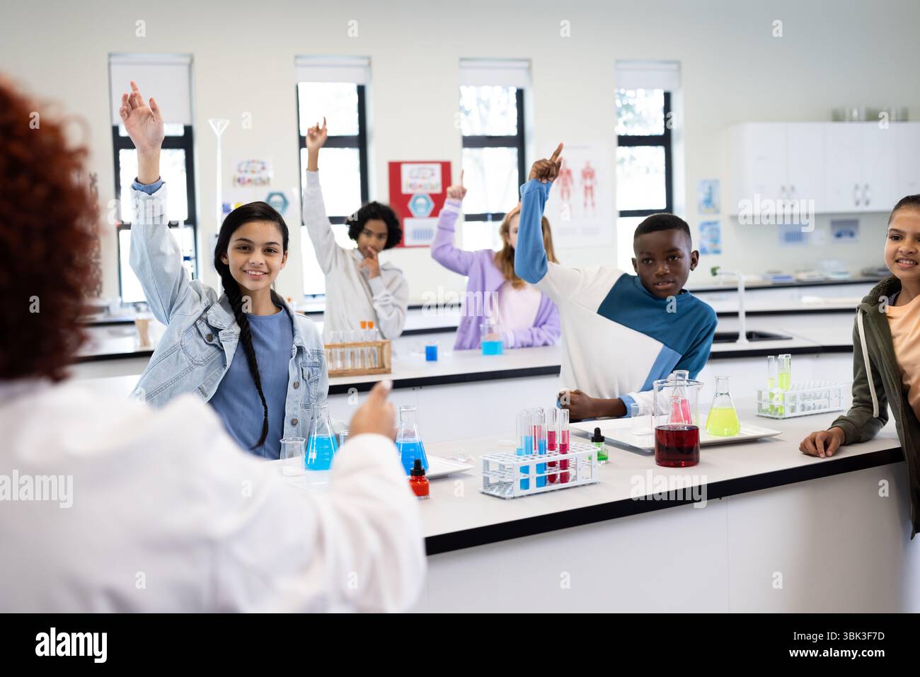 Studenti diversi che alzano la mano in classe di scienze, insegnante femminile ed esperimenti, a scuola Foto Stock