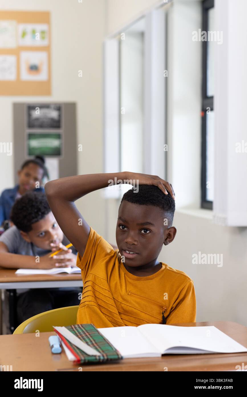 In classe, ragazzo afroamericano che alza la mano mentre i compagni di classe si concentrano sul loro lavoro, a scuola Foto Stock