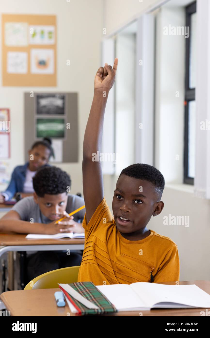 In classe, ragazzo afroamericano che alza la mano con impazienza mentre i compagni di classe si concentrano sul lavoro, a scuola Foto Stock