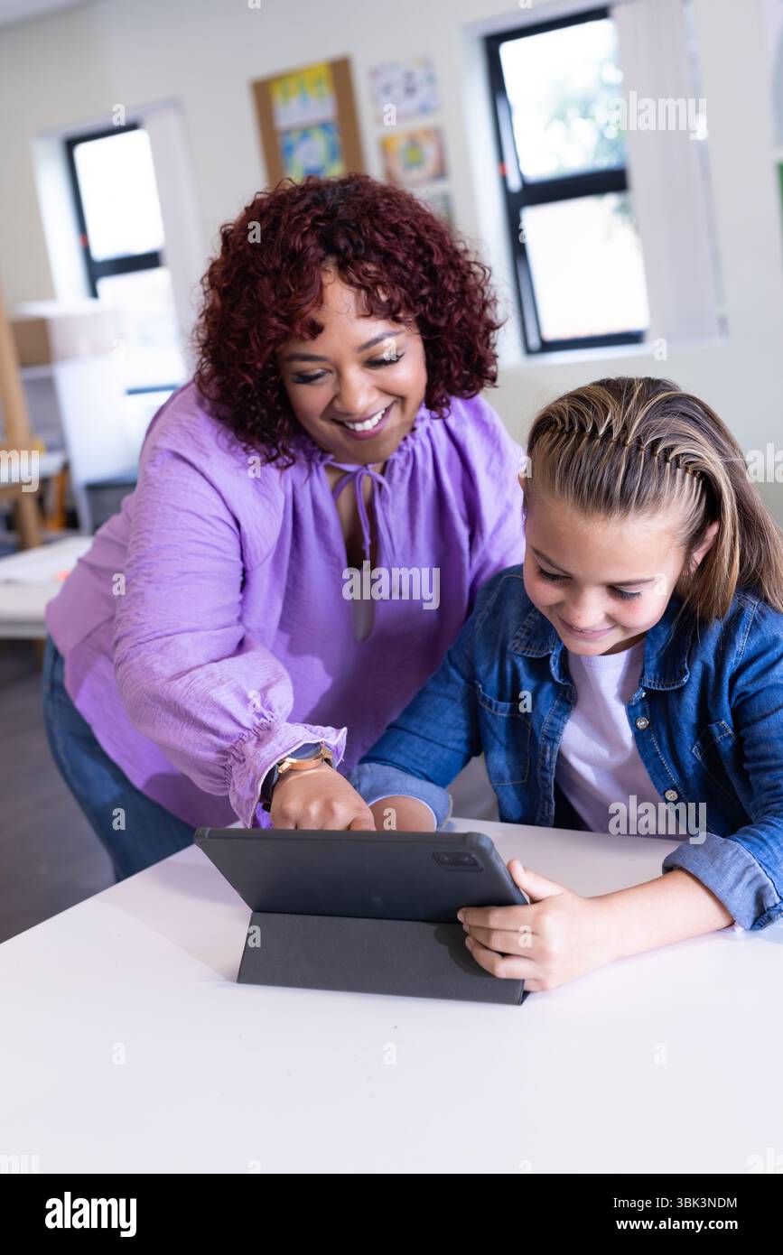 Insegnante donna che aiuta una ragazza con un tablet in classe, sorridente e impegnata, a scuola Foto Stock