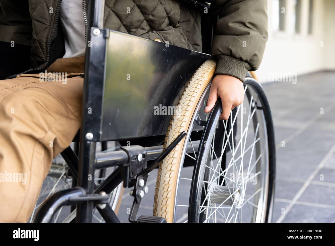 Ragazzo afroamericano in sedia a rotelle che va avanti, indossa una giacca calda e pantaloni marroni, a scuola Foto Stock