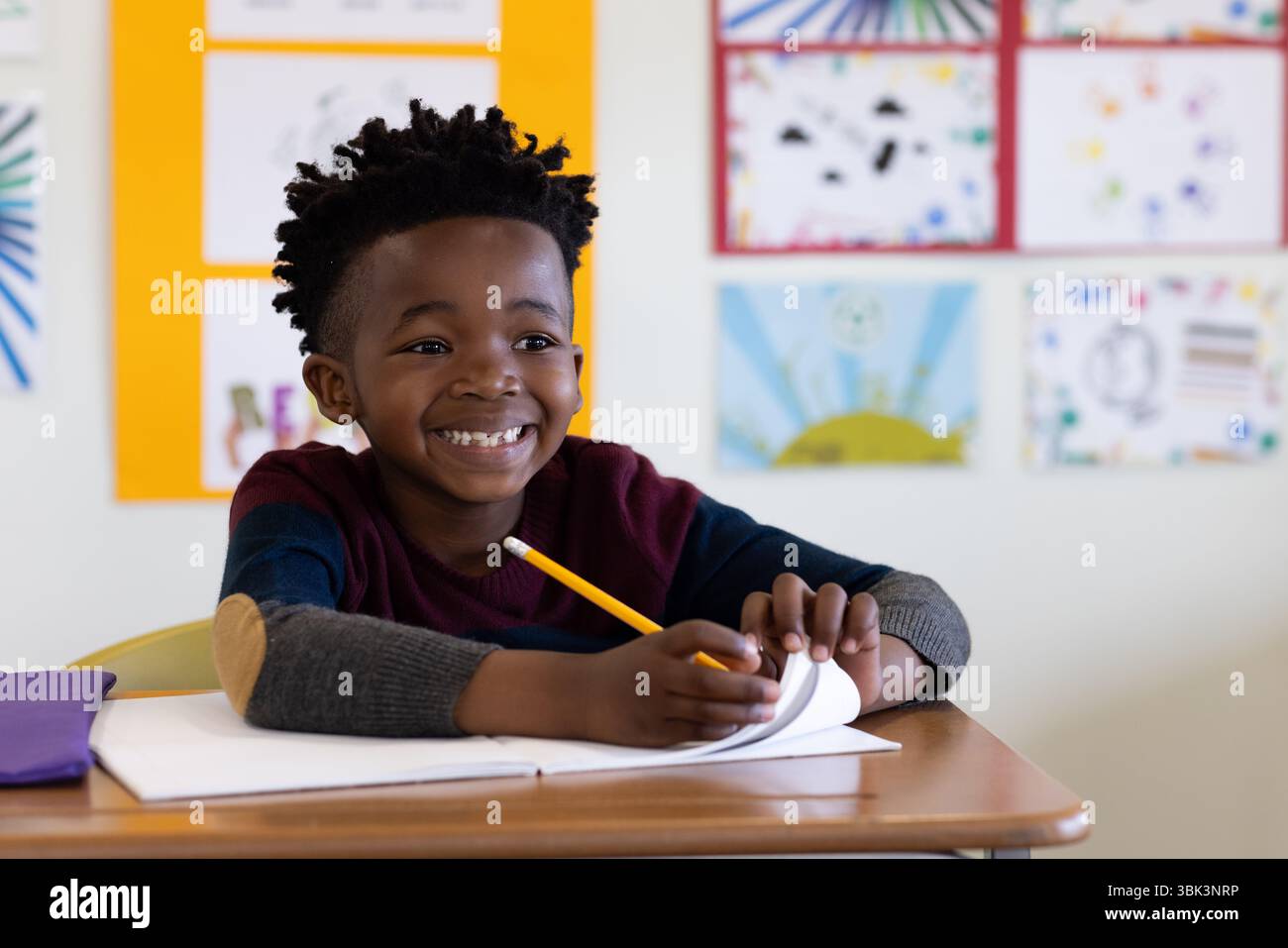 Sorridente ragazzo afro-americano a scuola che scrive con matita alla scrivania e si diverte ad attività in classe Foto Stock