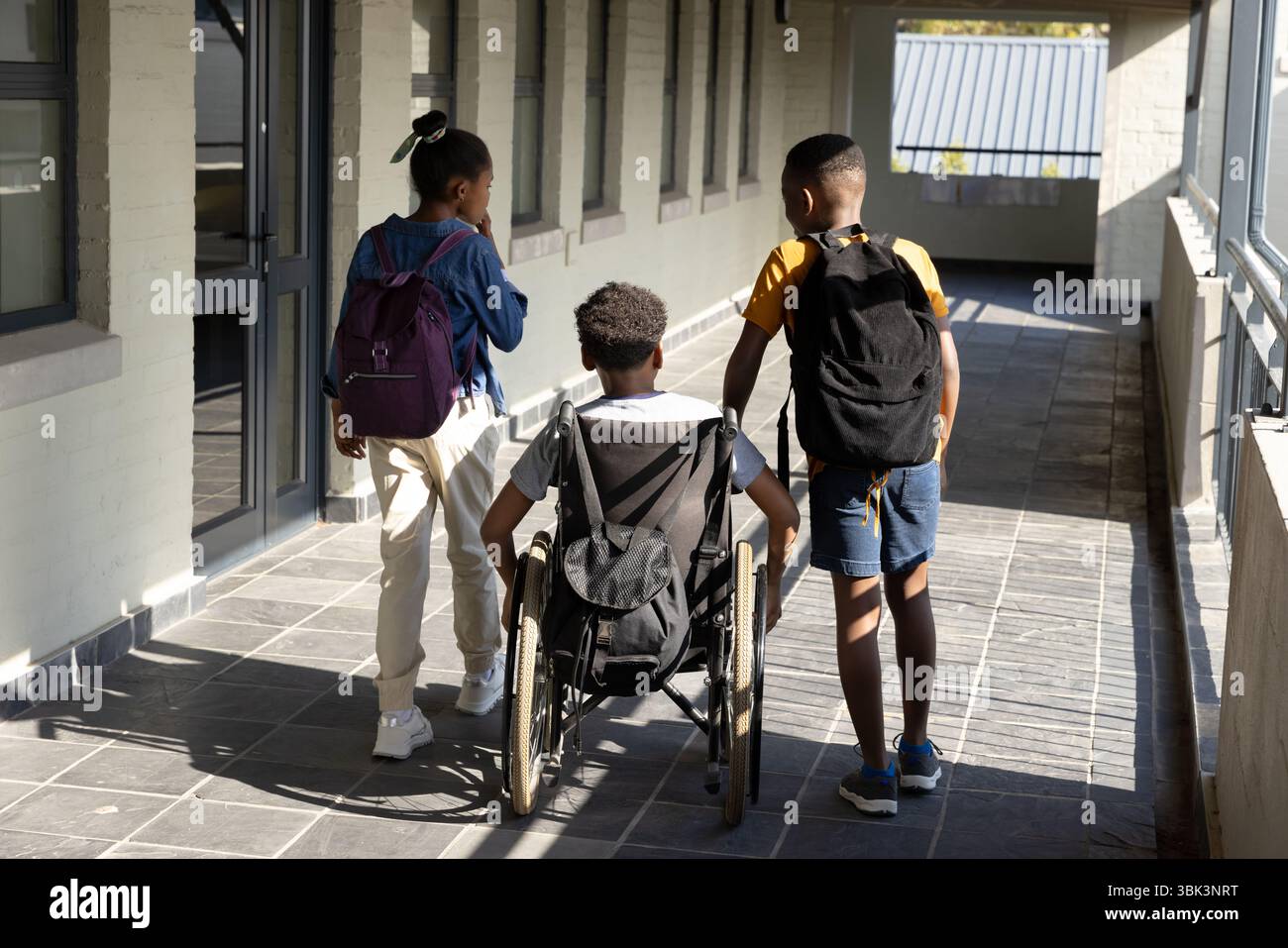 Studenti diversi che camminano insieme nel corridoio della scuola, sostenendo gli amici in sedia a rotelle Foto Stock