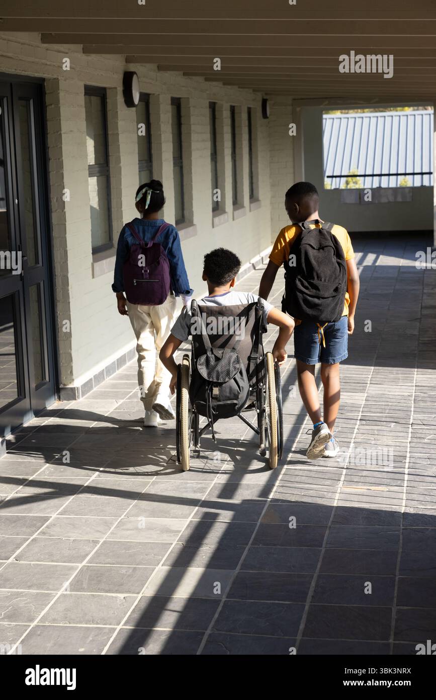 Studenti diversi che camminano insieme nel corridoio della scuola, sostenendo gli amici in sedia a rotelle Foto Stock