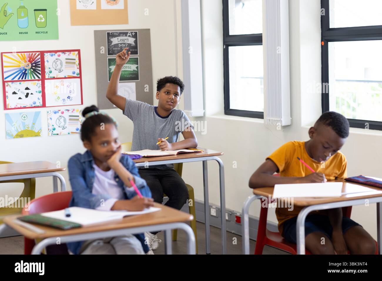 In classe, ragazzo che alza la mano mentre i compagni di classe scrivono con attenzione sui quaderni, a scuola Foto Stock