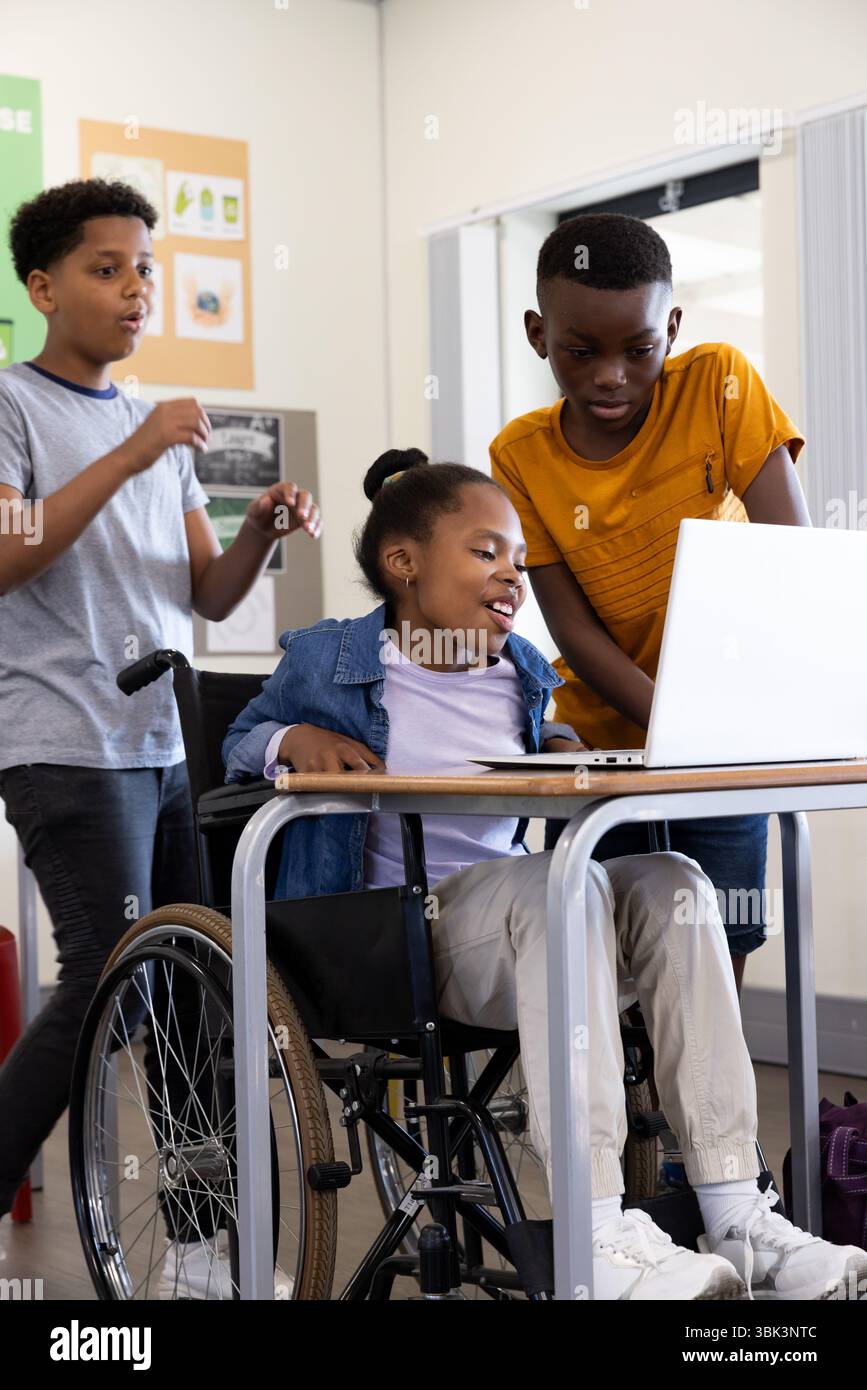 Studenti diversi che collaborano con un laptop in classe, ragazza in sedia a rotelle sorridente, a scuola Foto Stock