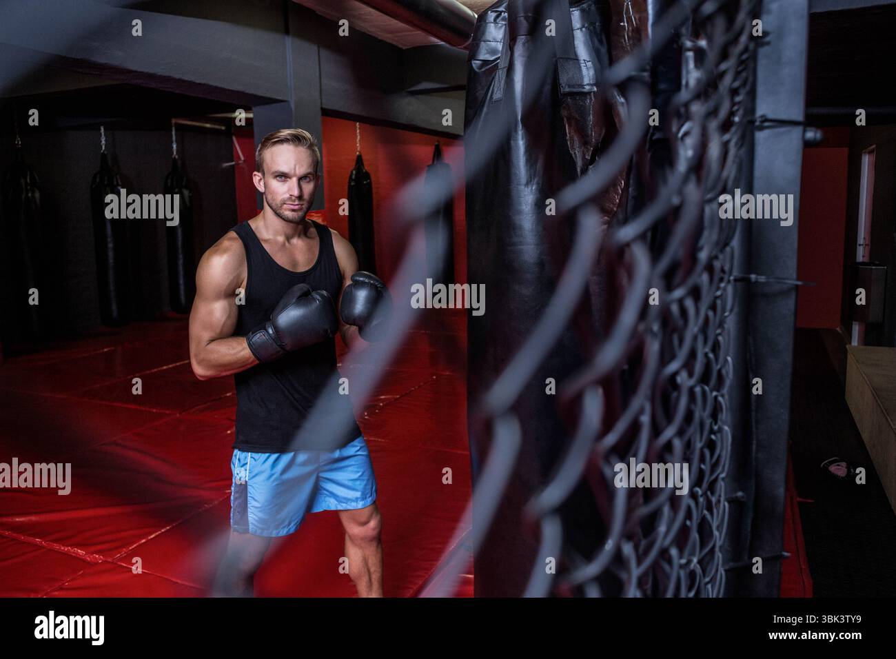 L'atleta maschile tira pugni contro una borsa pesante sul pavimento imbottito rosso in palestra con guanti da boxe Foto Stock