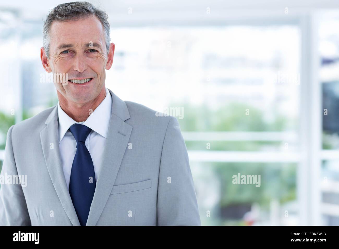 Uomo anziano in piedi in ufficio vicino a grandi finestre, indossa una tuta grigia, sorride e guarda la macchina fotografica Foto Stock
