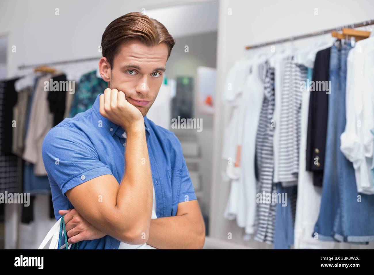 Uomo appoggiato sulla testa del pugno che tiene la borsa della spesa all'interno del negozio di abbigliamento tra scaffali, specchio, spazio per le copie Foto Stock