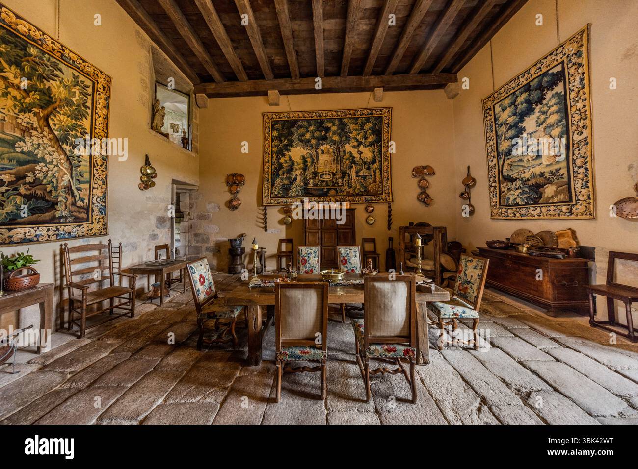 Tre grandi arazzi paesaggistici (verdures) adornano una sala nello Château de Boussac, dipartimento di Creuse, Nouvelle-Aquitaine, Francia. Mostrano alberi, parchi e uccelli e sono appesi alle pareti chiare, incorniciati da bordi con fiori e ornamenti. Un lungo tavolo di legno con candelabri si trova al centro, circondato da sedie con rivestimenti floreali. Oggetti in rame sono appesi alle pareti, una lunga cassa di legno con ciotole si trova a destra e un armadio di legno è posizionato sul retro. Il pavimento è ricoperto da grandi lastre di pietra e il soffitto ha travi di legno scuro. Il castello è una fortezza medievale con una ricca hi Foto Stock