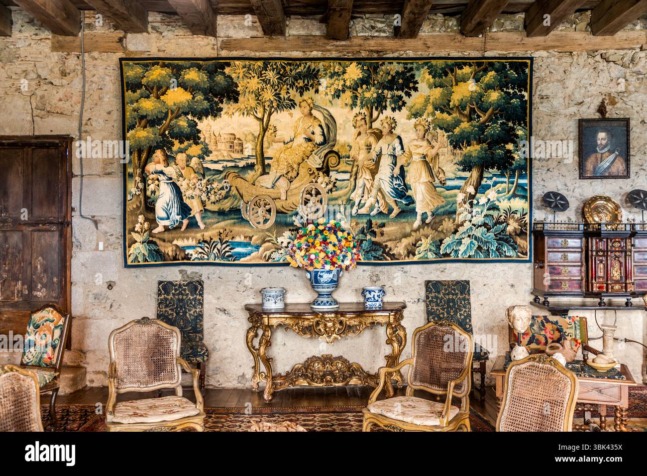 Un grande arazzo è appeso in una stanza dello Château de Boussac, dipartimento di Creuse, Nouvelle-Aquitaine, Francia, sopra un tavolo console dorato e intarsiato. Raffigura una donna su un carro con ruote a raggi, accompagnata da donne in abiti fluenti e bambini in un paesaggio boscoso con un castello sullo sfondo. Un bouquet colorato in un vaso blu e bianco si trova sulla console tra due vasi blu e bianchi. Sedie con schienale in vimini sono posizionate davanti, e sedie imbottite si appoggiano contro il muro. A destra, un ritratto di un uomo con una collana a balze è appeso sopra un armadio con cassetti sormontati da piastre di ottone. Sul l Foto Stock