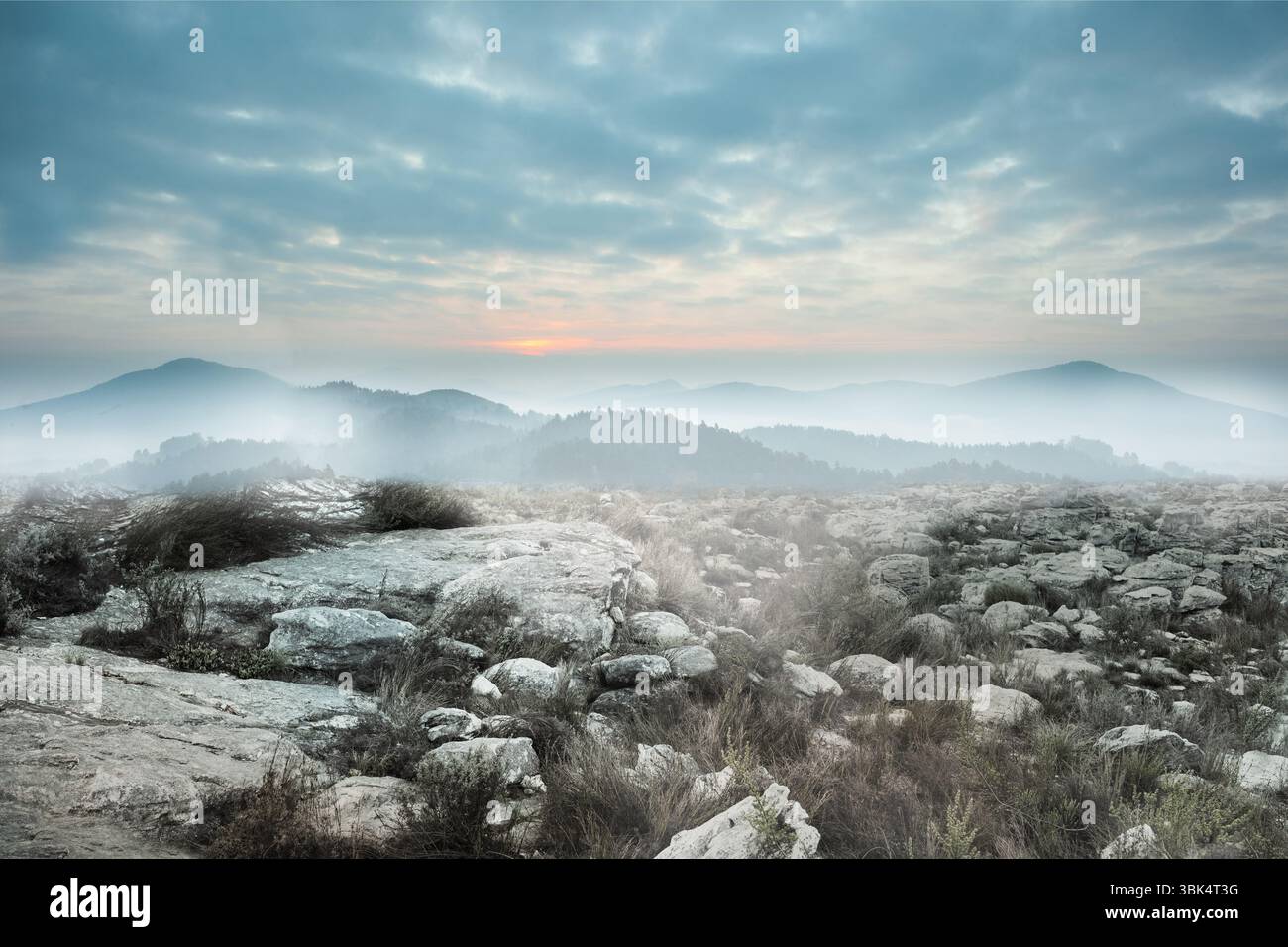 L'altopiano roccioso si estende su un design piatto, mostrando massi pallidi, colline nebbiose e bagliore arancione Foto Stock